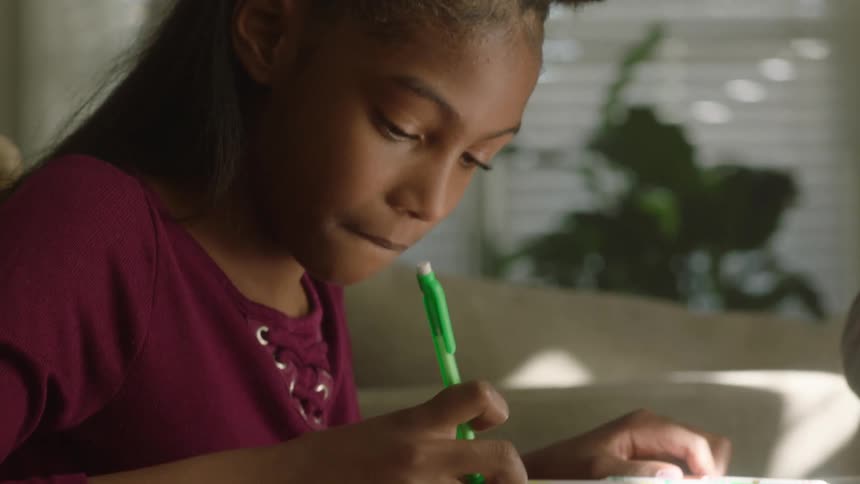 A black female child writing on a book with a pen while seated on a chair indoor