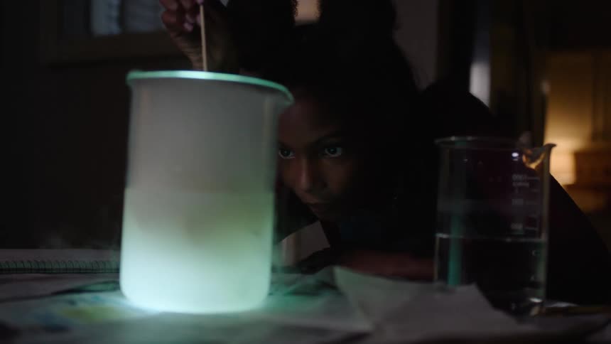 A female child studying the liquid material inside a cone placed on the table in a dark room