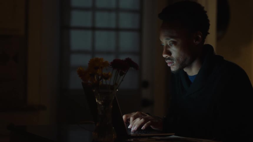 A man is sitting a desk in a dark room typing on a laptop computer