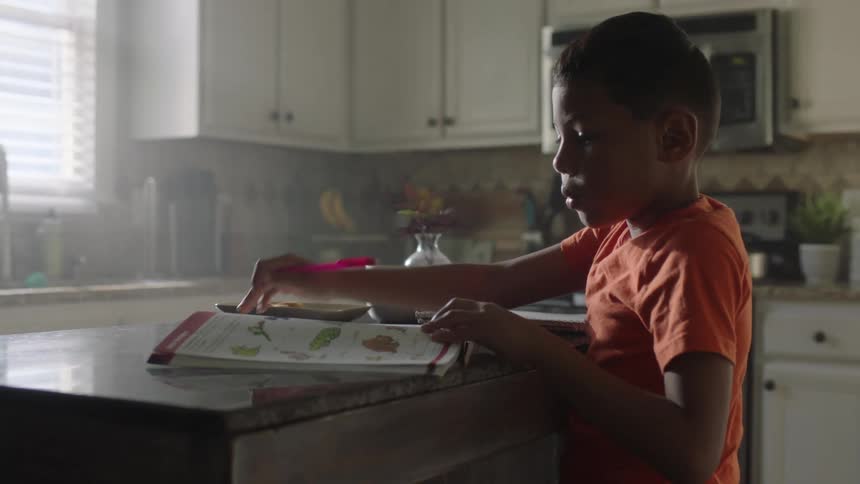 A little boy is turning the page of a book and reading it while holding a pink pen in his hand