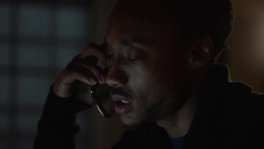 A black man in a dark room talking on his phone while working on his laptop