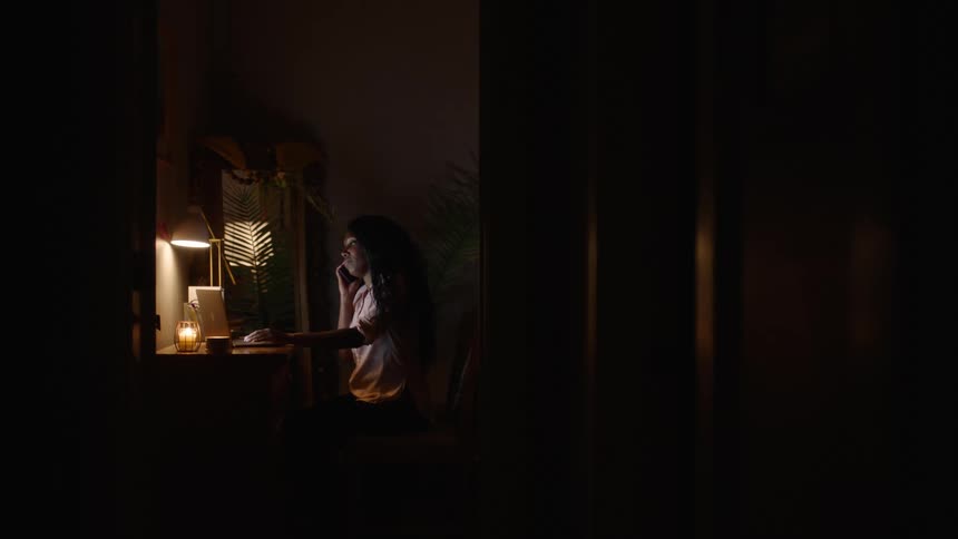 A man sitting in a dark room operating his computer and talking on his phone