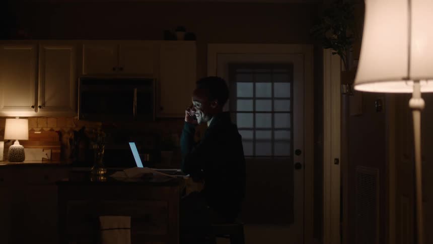 A young man is sitting a desk in a dark room looking at a laptop computer and laughs when he answers his phone