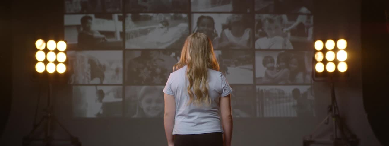 A girl turns her back and looks at a screen with several videos of people