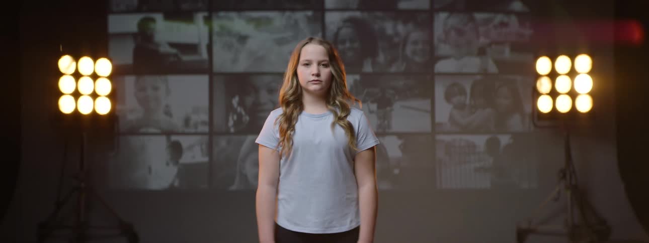 Portrait of a young blonde teenage girl standing still with a screen behind her playing footage of children