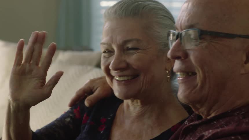 An elderly couple sitting together on the couch waving into a cell phone