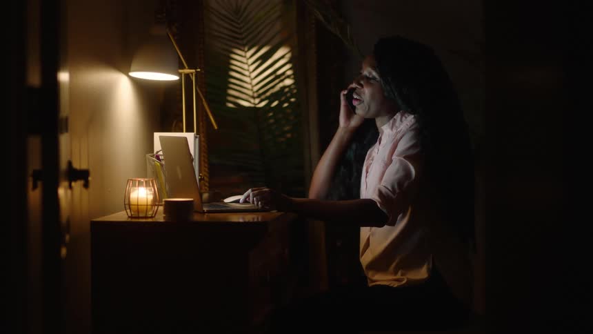 A woman is sitting in a dark room in front of a laptop computer as she talks on the phone