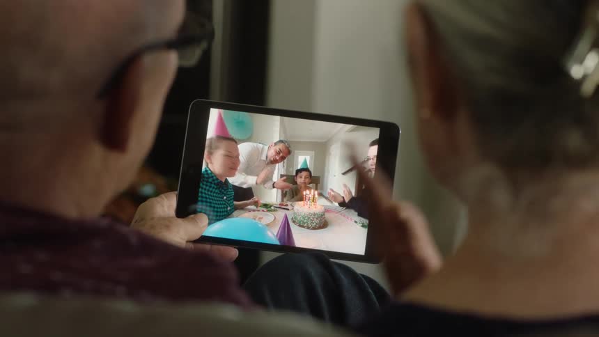 A birthday party live streams on a tablet between the backs of a mans and a womans head who are sitting beside each other while the man who is holding the tablet sings and the woman claps her hands