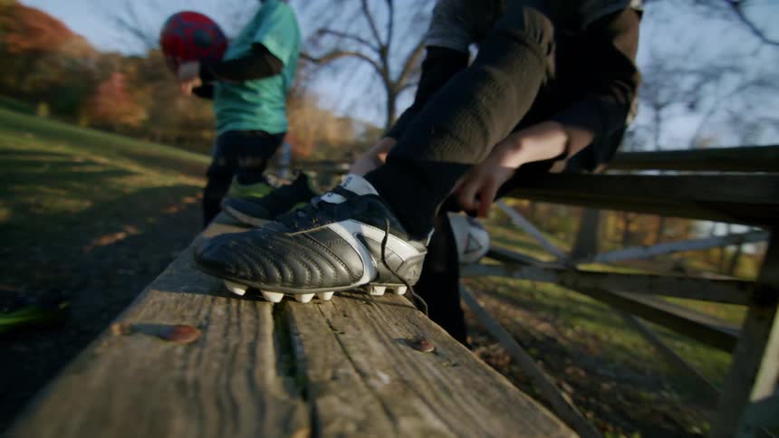 One boy juggles a soccer ball as another boy puts a cleat on his left foot