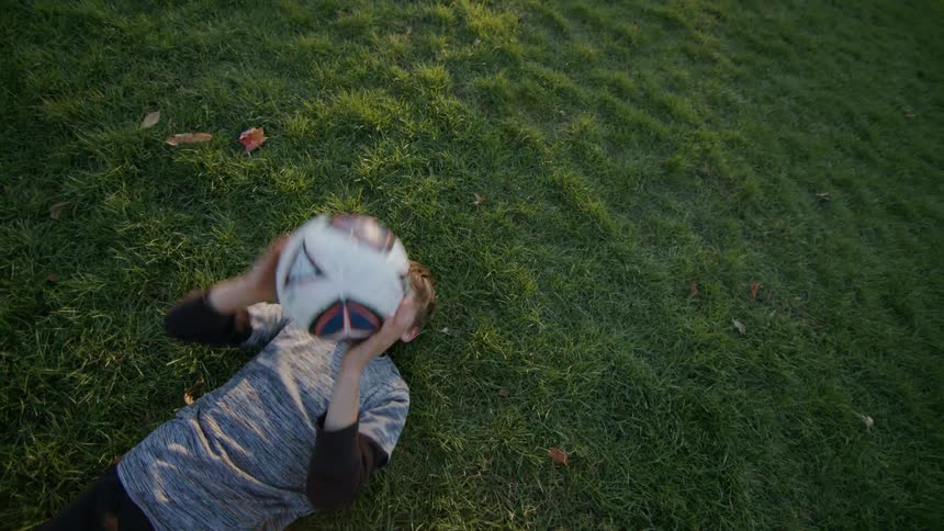 A young boy throwing a ball up while laying on the grass outdoor
