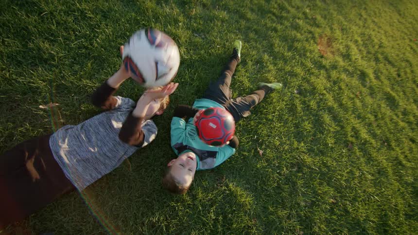 Two boys are laying down on the soccer field and throwing soccer balls up in the air then catching them