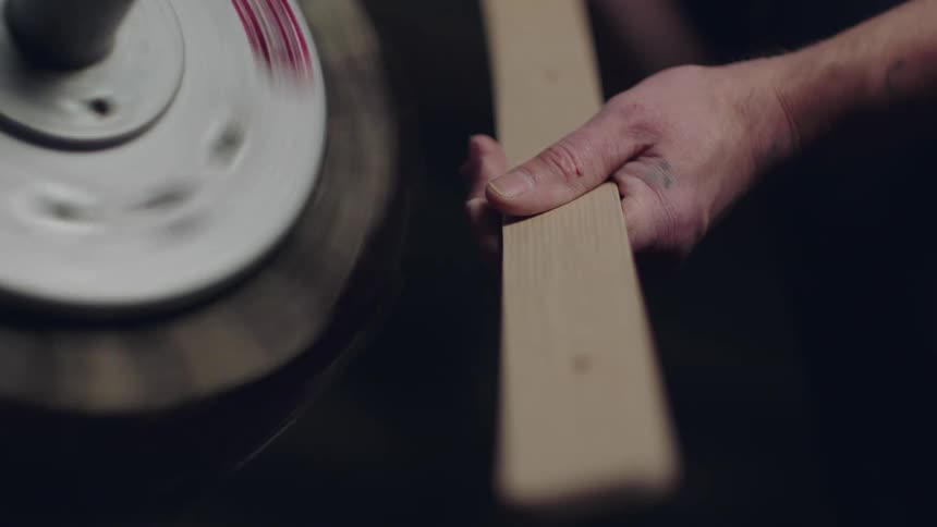 Thin wood edge boards stacked together move through a persons hand beside a wheel with a black buffing edge that is spinning quickly around a post
