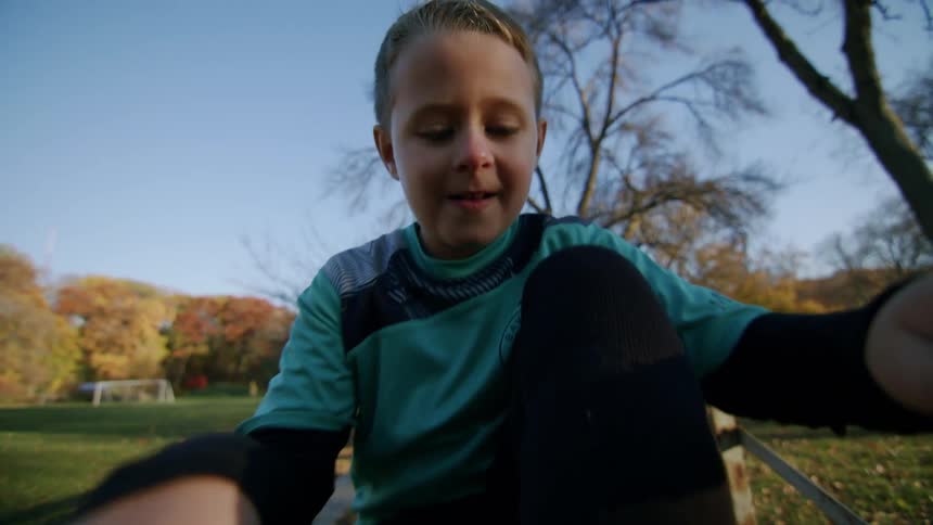 A young boy ties the laces of his soccer cleats while on a wooden bench