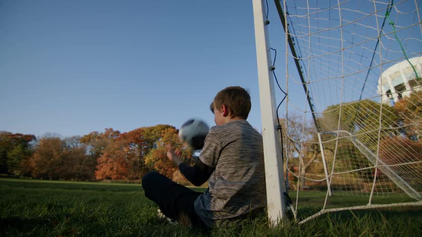 A kid playing with a ball while sitting by the goal post in a football field