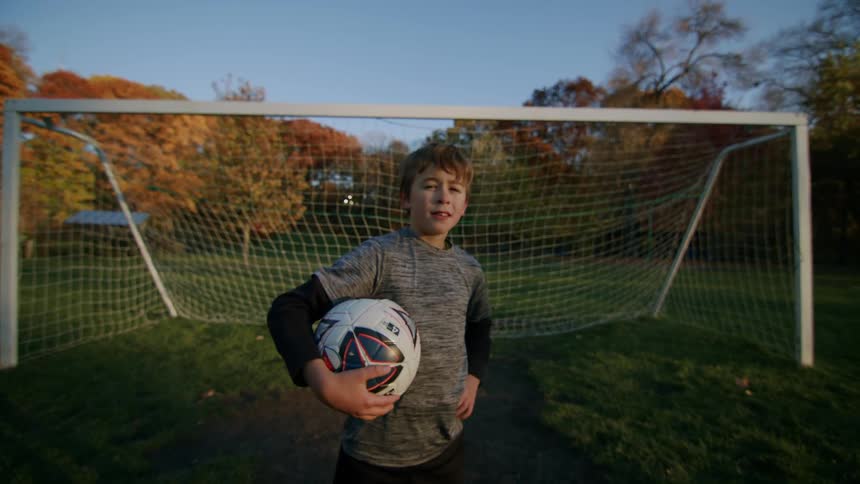 A boy confidently posing for the camera in front of the soccer goal holding a ball