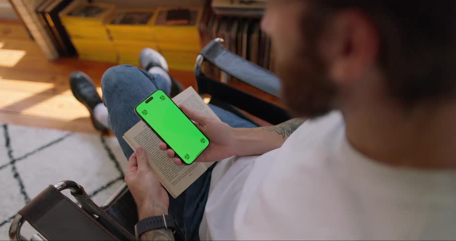 A man holding a mobile phone on the right hand and a book on the left hand while seated on a chair indoor at midday