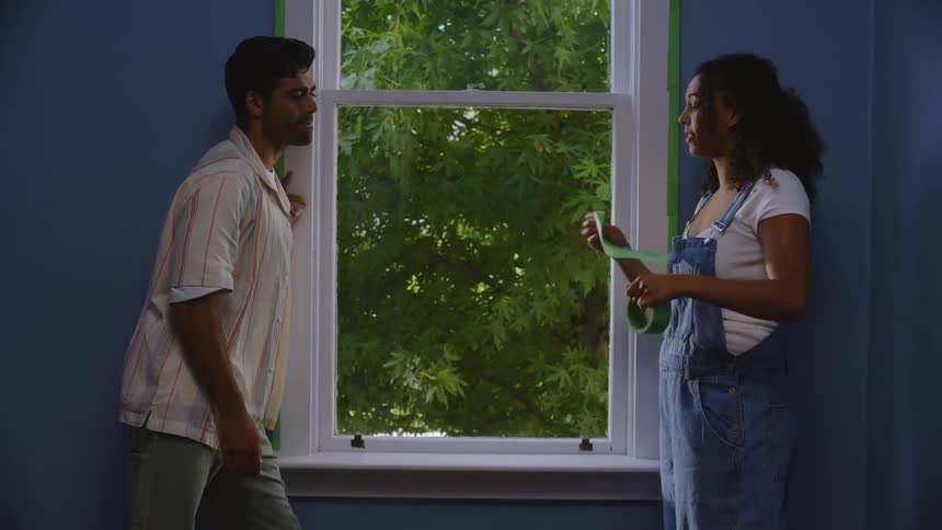 A woman puts painters tape around a window while a man helps adjust it before they both sit on the window sill with the mans arm around the woman