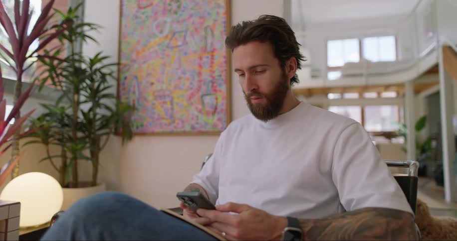 A beard man sitting indoor while holding a book and pressing his phone