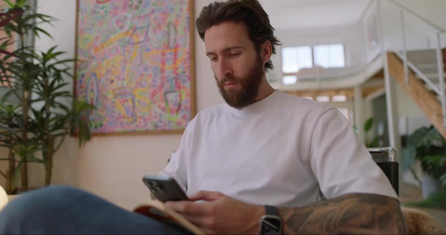 A bearded man in a white tshirt jeans and with a tattoo on his arm sits in an armchair using an iphone smartphone