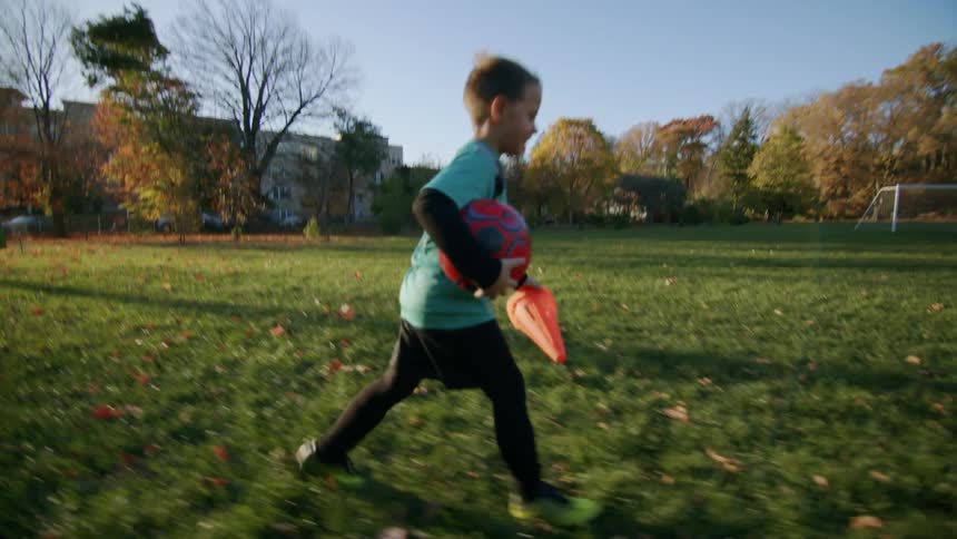A little boy is running across a grassy field holding a soccer ball in his arm