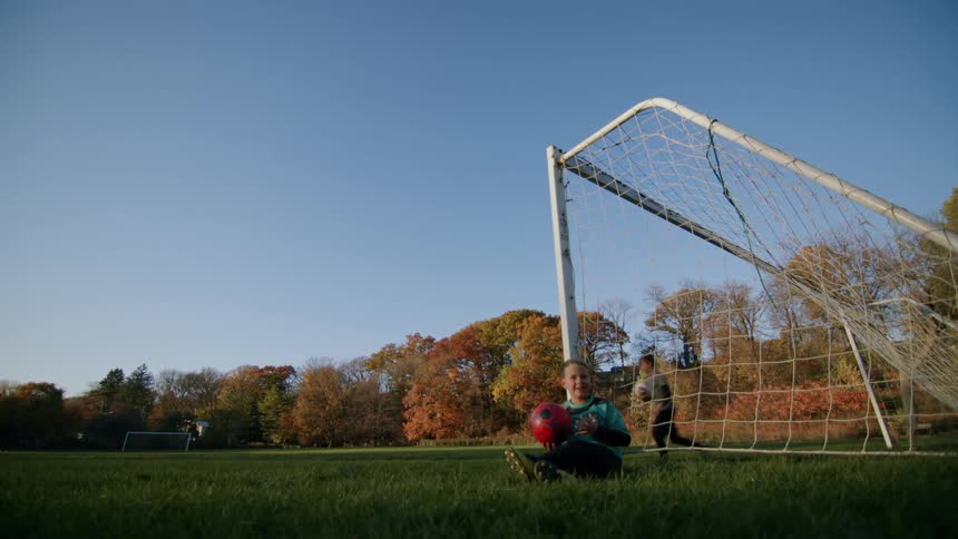 Boy leans against soccer goal post while brother practices with soccer ball during the fall