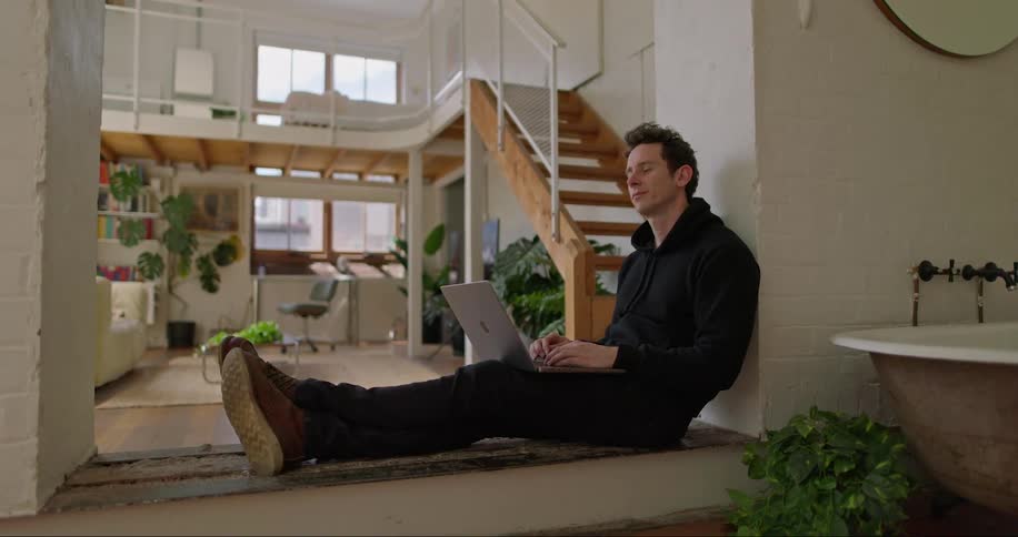 Man types on a laptop while seated on the floor indoor at midday