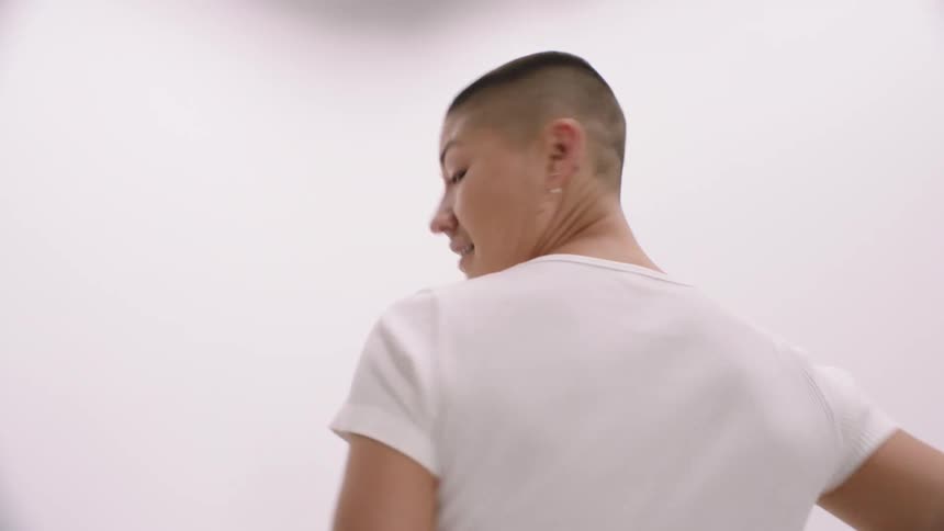 Woman with a buzz cut models jeans by walking into a white corner turning around and walking back again