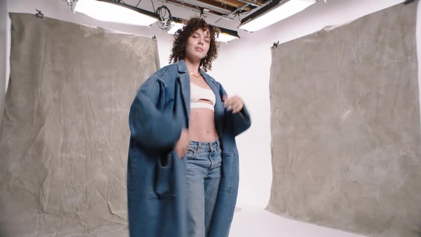A woman dances around in a small room modeling clothes and removing a jacket