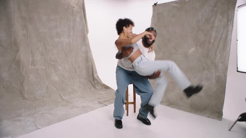 A guy in a white shirt and jeans joyfully lifts a woman with curly hair both wearing crop tops and jeans amidst the studio backdrop sharing a laugh quirky white cyc