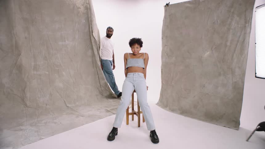 A woman on a bench prepares for a white cyc photo shoot studio as man comes from behind to surprise her quirky
