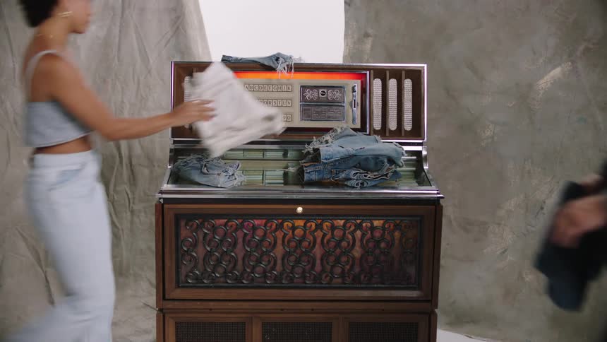 Multiple people place denim clothes on jukebox in photography studio before a man selects a song white cyc