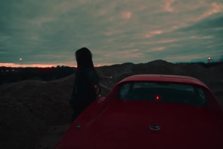 A woman leaning against a red car observing the landscape during dusk