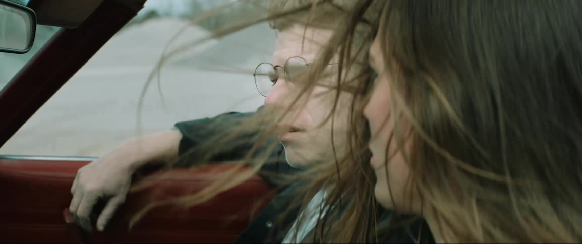 A woman with a pained expression drives a sports car down a dirt road sharing a moment of sorrow with a man beside her as they both shed tears together
