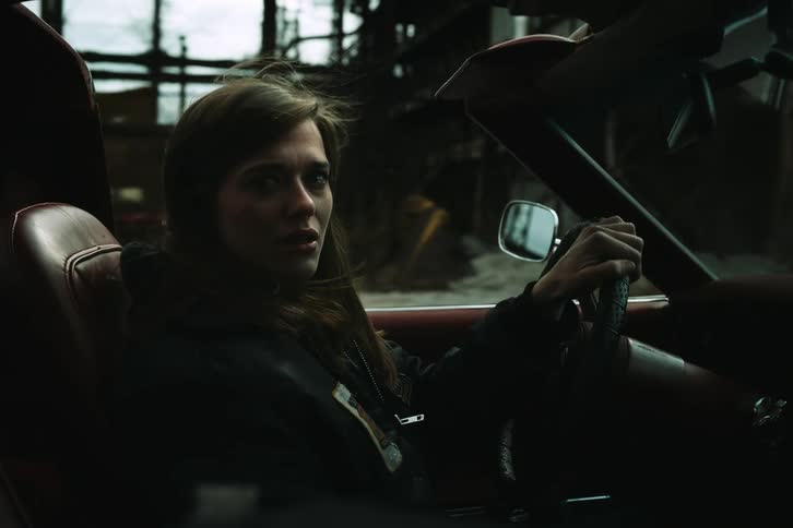 A woman with brown hair drives a sports car down a dirt road tears streaming down her face expressing sadness