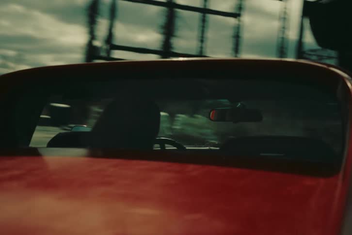 We see from behind the driver that someone is driving a red red car along a highway past buildings and construction sites other vehicles sky is blue sunny