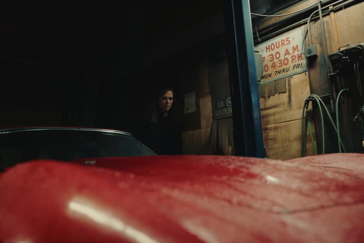 A woman with brown wavy hair and a black coat steps out of a broken red sports car and checks the engine in the garage
