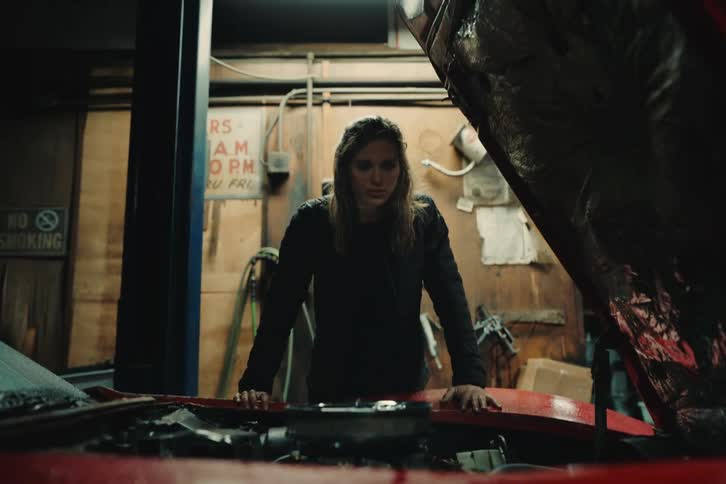A woman with brown hair and a black coat inspects the engine of a red sports car