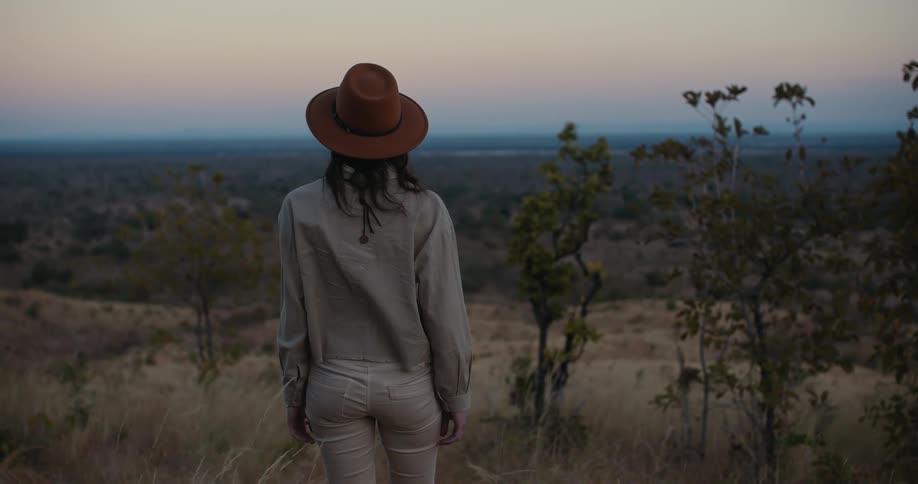 A woman in a safari hat moves forward through tall brown grass turns her head looking side to side out at the african plains at dusk