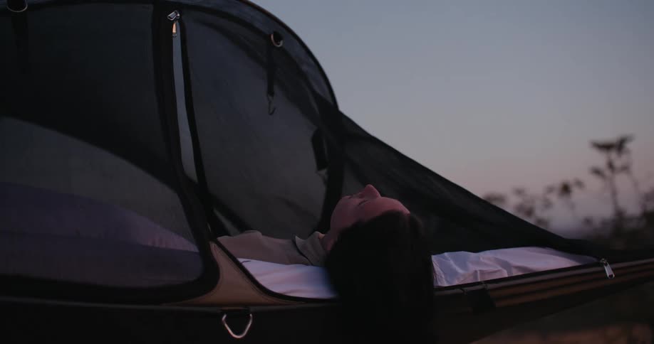 A woman lying in a suspended tent looking at the sky at sunset