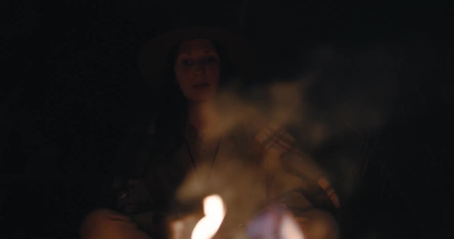 Woman sits near smoking campfire at night and looks around