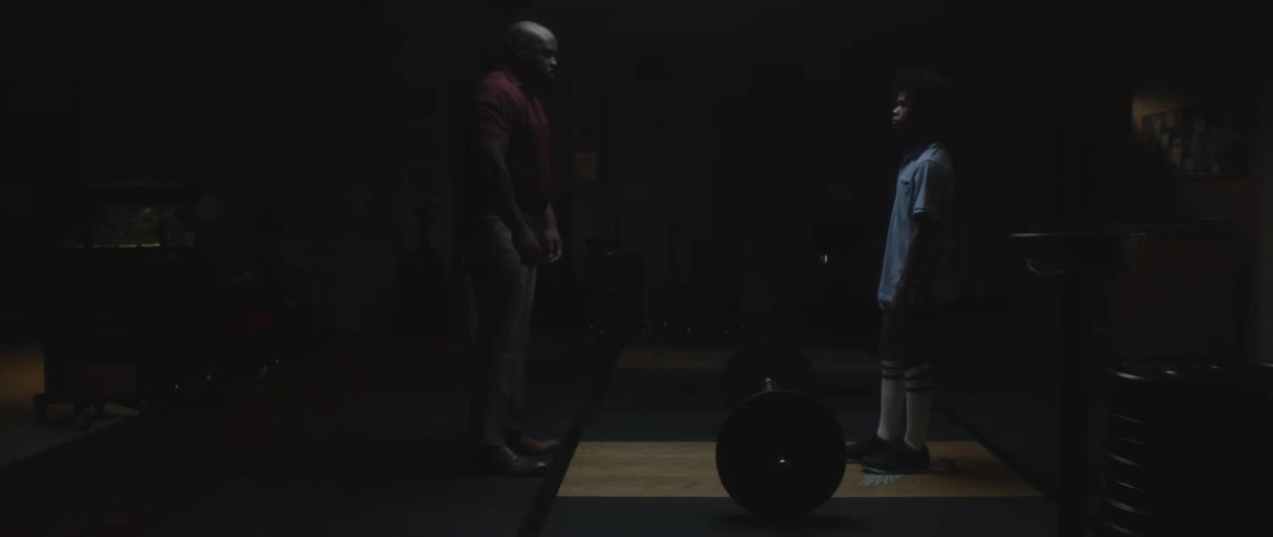 Adult black man and a teenager black boy standing up facing each other in a hostile manner indoors at a small gym
