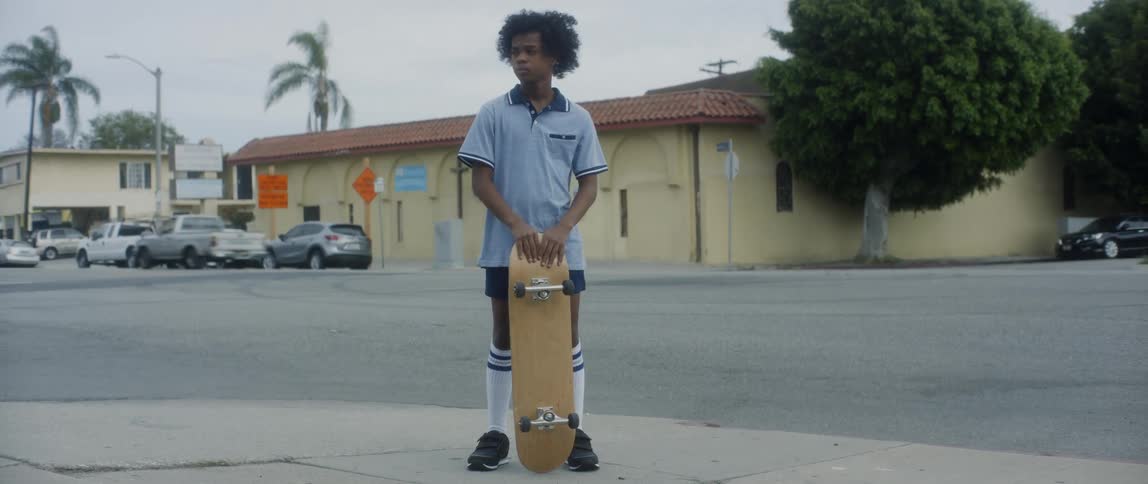 A kid holding a skateboard while standing on the side of a busy road