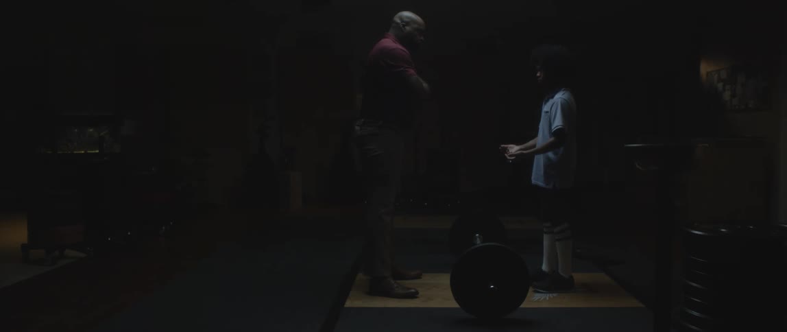 Man coaches youth in lifting weight bar in dimly lit room
