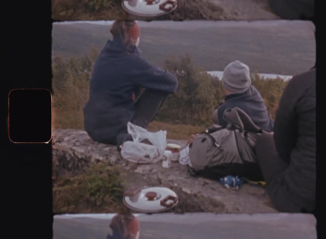 Three campers sit on rock slab looking over lake bordered by forest and mountains while a meal cooks