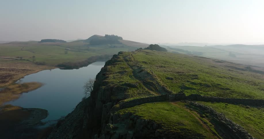Aerial of a rock wall on the edge of a green covered highland cliff beside a clear lake with farmland opposite the cliff