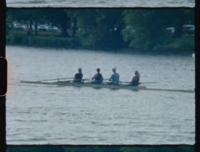 Four people in a canoe row across a lake in a recreation area moving their oars in sync