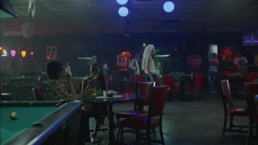 A woman is slowly walking into an active pool hall with people drinking and playing pool