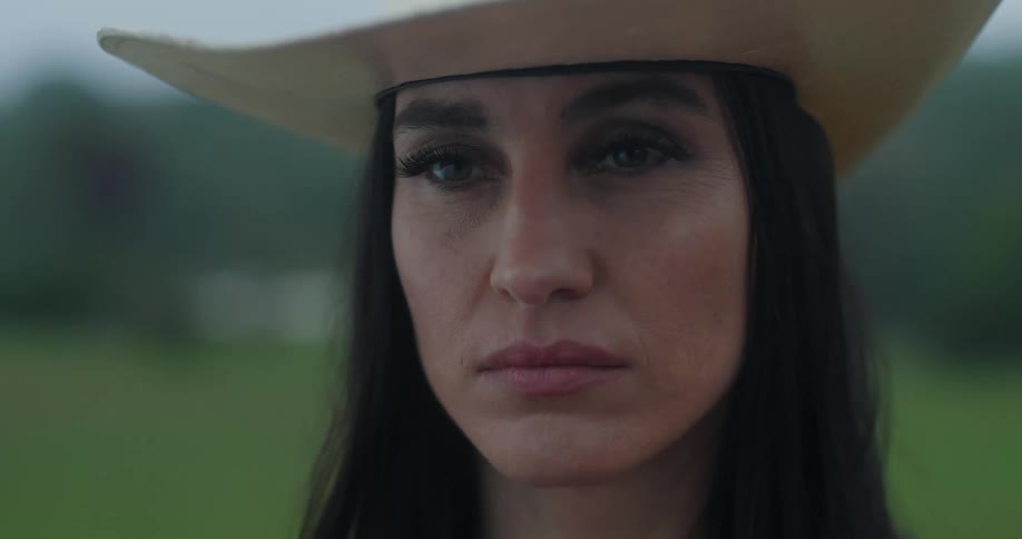 Closeup of a woman with a solemn expression wearing a cowboy hat with a blurred green field in the background