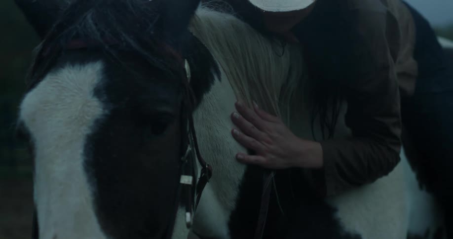 A woman sitting on a horse with her torso bent forward laying on its neck brushes down its mane while rain falls on them from a clouded sky