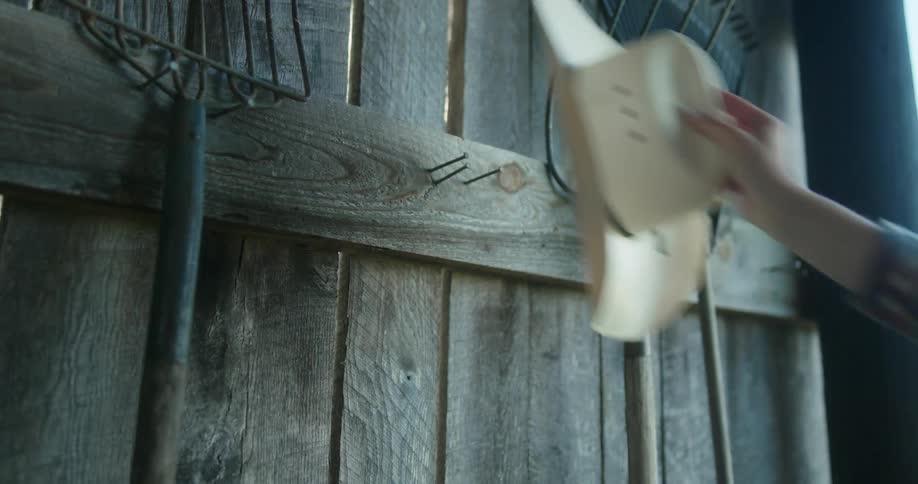 Person picks up cowboy hat from wall next to pitchfork then hangs it again
